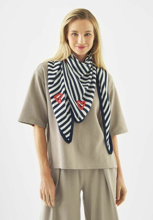 Vrouw gekleed in een beige loszittende outfit en een marineblauw-wit gestreepte sjaal met rode hartmotieven, glimlachend naar de camera tegen een witte achtergrond.