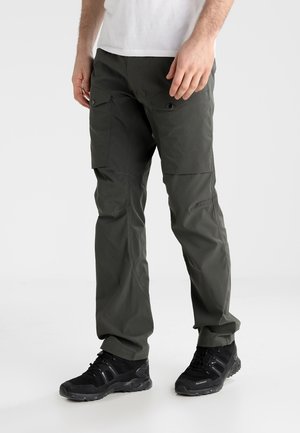 Mann trägt dunkelgraue Cargohosen mit geknöpften Oberschenkeltaschen und schwarzen Sportschuhen vor einfarbigem Hintergrund.
