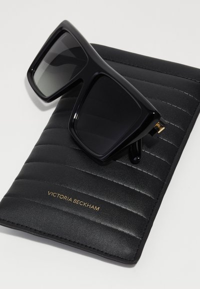 Crne prevelike sunčane naočale s tamnim staklima odmaraju se na teksturiranoj crnoj futroli, ugraviranoj s "Victoria Beckham" u zlatnoj boji.