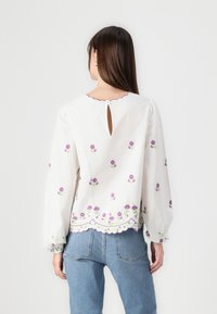 Blusa bianca con ricami floreali viola, orlo a scallop e dettaglio a chiave sul collo. Maniche lunghe a sbuffo con polsini elastici. Vestibilità casual.