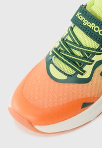 Sport sneaker with an orange mesh upper, green accents, and textured detail. Features a lace-up design and a cushioned white midsole.