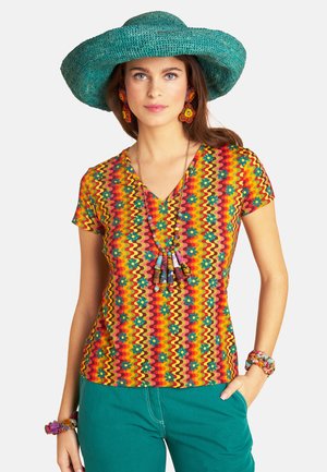 Woman wearing a colorful patterned V-neck shirt, teal wide-brim hat, matching teal pants, floral earrings, and layered beaded necklace.