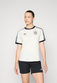 Jaśkofioletowa koszulka sportowa z granatowymi akcentami, z okrągłym dekoltem, krótkimi rękawami i naszywką na piersi. W połączeniu z czarnymi szortami.