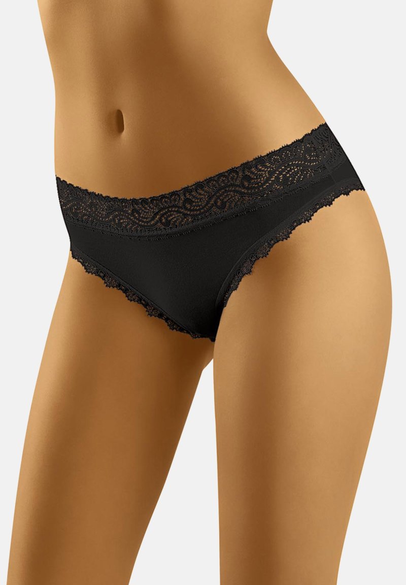 Femme portant des sous-vêtements de style bikini ornés de dentelle noire, mettant en avant son ventre et le haut de ses cuisses sur un fond uni.