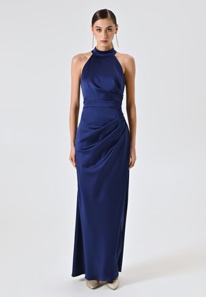 Robe maxi satin marine à col halter, taille ajustée, détail drapé sur le côté et texture lisse et fluide. Association avec des talons de couleur claire.