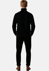 Schwarzer Rollkragenpullover kombiniert mit schwarzen Hosen. Die Rückansicht zeigt ein figurnahes Design mit Rippdetails an den Bündchen und am Saum. Braune Schuhe.