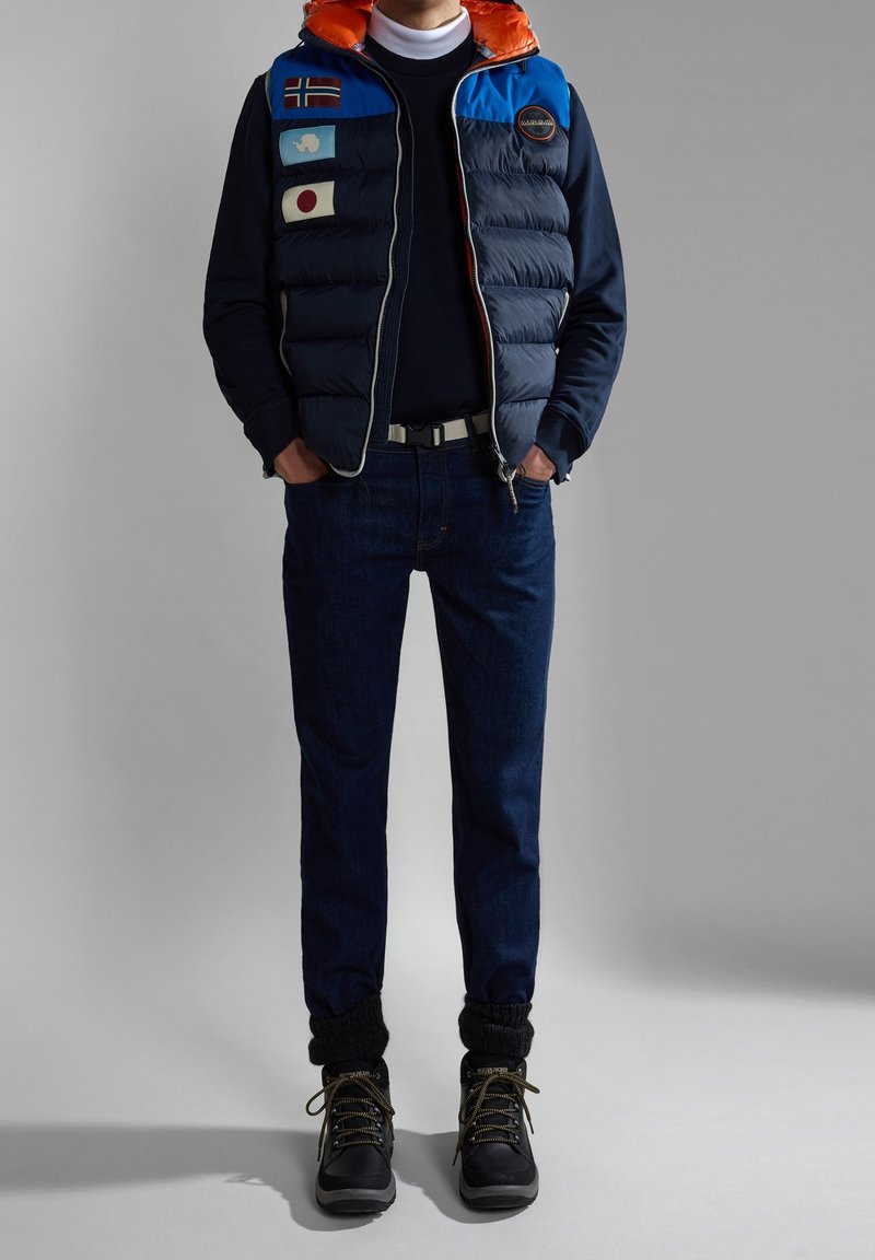 Gilet matelassé bleu marine avec capuche bleue et orange, orné de drapeaux internationaux. Porté avec un jean en denim foncé et des bottes de randonnée noires.