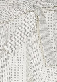 Bílá látka s vertikálními černými pruhy, která má detail vázaného pásku. Materiál vypadá hladce s mírnou strukturou.