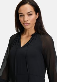 Schwarze Bluse mit V-Ausschnitt, gerafftem Ausschnitt und langen transparenten Ärmeln. Sie hat eine leichte Textur und einen lockeren Schnitt, der sich gut zum Layering eignet.