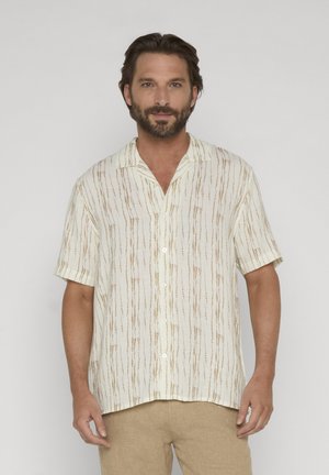 Uomo con barba che indossa una camicia bianca a maniche corte con bottoni e strisce verticali marroni, e pantaloni beige, in piedi davanti a uno sfondo semplice.