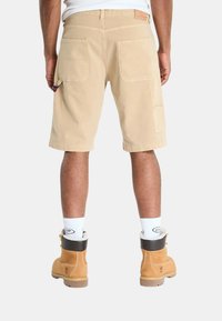 Pantaloni cargo beige con due tasche posteriori e una tasca laterale per attrezzi. Indossati con stivali da lavoro marroni e calzini bianchi fino alla caviglia. Design a gamba dritta.