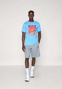Lysblå T-shirt med rødt Nike-logo, parret med grå sportsbukser. Modellen har hvide sneakers og hvide sokker med et sort logo på.