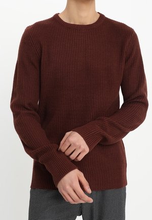 Person trägt einen braunen gerippten Langarmpullover und schwarz-weiße karierte Hose, die Hände vor sich verschränkt.
