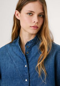Denimhemd mit hohem Kragen, gerafftem Ausschnitt, Knopfverschluss und kontrastierenden Metallbeschlägen; verfügt über eine einfarbige blaue Farbe und eine sanfte Textur.