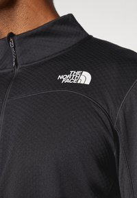 Haut à manches longues zippé noir en tissu texturé, avec un col montant et un logo blanc "The North Face" sur l'épaule gauche.