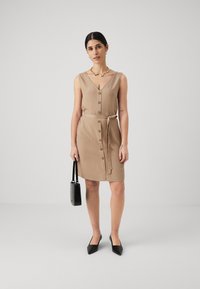 Beige ermeløs kjole med V-hals, frontknapper og knyttet midje. Kombinert med en liten svart veske og spiss-sko i svart.