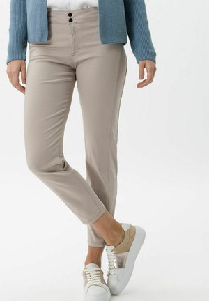 Persoon draagt beige slim-fit broek, wit-beige veter sneakers en een blauwe gebreide trui, staand met gekruiste benen.