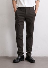 Man wearing black slim-fit pants and black leather loafers, hands in pockets, standing against a light textured wall.