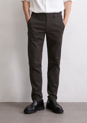 Man in zwart slim-fit broek en zwarte leren instappers, met handen in de zakken, staande tegen een lichte gestructureerde muur.