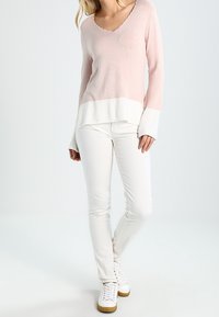 Pull en maille rose clair avec un col en V et un ourlet blanc, associé à un pantalon blanc ajusté et des baskets blanches. Texture lisse et design décontracté.