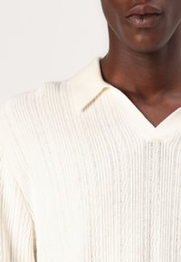 Polo em malha marfim com decote em V, apresentando textura ribbed vertical e um brilho sutil, feito de um tecido suave com um design de colarinho.