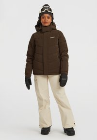 Bruine pufferjack met een hoge kraag, ritszakken en logo. Gecombineerd met crèmekleurige sneeuwbroeken en zwarte handschoenen, geschikt voor wintersport.