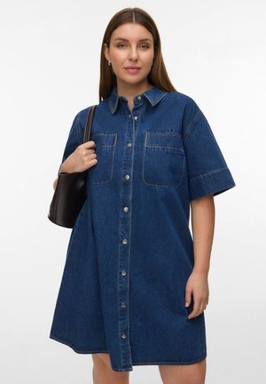 Femme portant une robe en jean bleue à manches courtes boutonnée, tenant un sac noir à l'épaule sur un fond blanc uni.