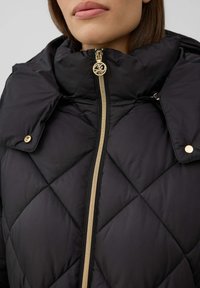 Femme portant une veste matelassée noire avec une fermeture éclair dorée et des boutons-pression, mettant l'accent sur le col montant et la partie supérieure de la poitrine.