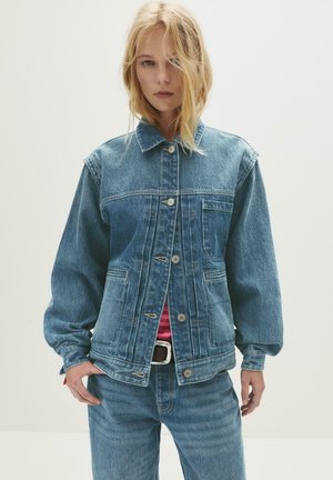 Vrouw met blond haar die een blauwe spijkerjack en jeans draagt, staande voor een effen lichte achtergrond.