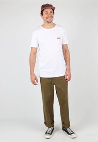 T-shirt blanc à manches courtes avec un petit motif rouge et blanc sur la poitrine, associé à un pantalon vert olive et des baskets noires.