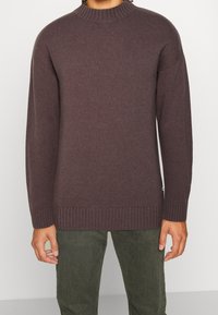 Dunkelbrauner Strickpullover mit einem hohen Rippkragen, überschnittenen Schultern und gerippten Manschetten, kombiniert mit olivgrünen Hosen.