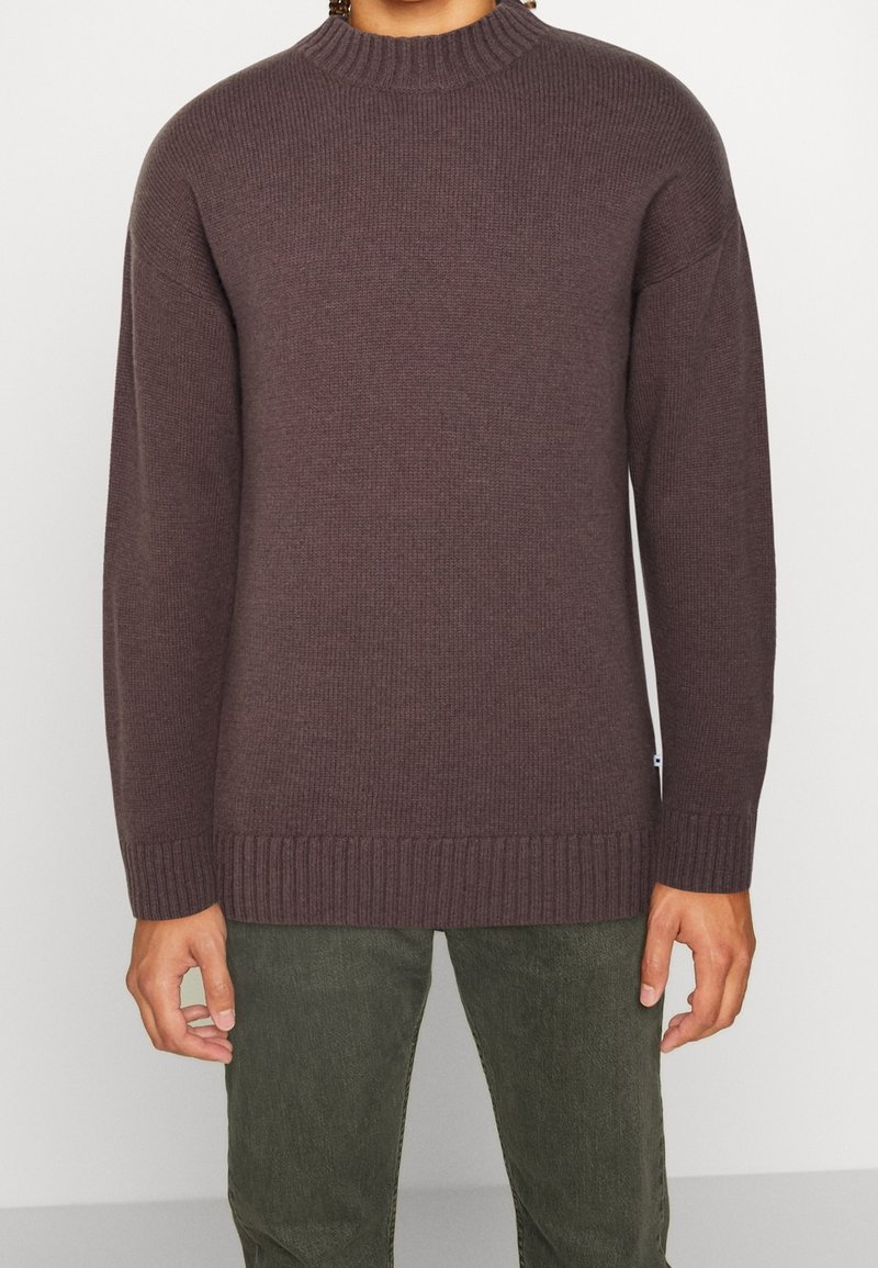 Dunkelbrauner Strickpullover mit einem hohen Rippkragen, überschnittenen Schultern und gerippten Manschetten, kombiniert mit olivgrünen Hosen.