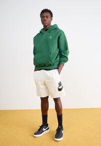 Zelená mikina s klokaním vreckom, čierne logo na hrudi, skombinovaná s krémovými šortkami s veľkým čiernym logom Nike, čierne tenisky a tmavé ponožky.