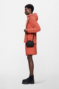 Un modelo con una chaqueta impermeable naranja y pantalones cortos, combinados con botas negras elegantes hasta el tobillo y un pequeño bolso negro de bandolera.