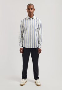 Gestreiftes Hemd aus leichtem Stoff mit vertikalen blauen und weißen Streifen; kombiniert mit dunkler Hose und weißen Sneakers.