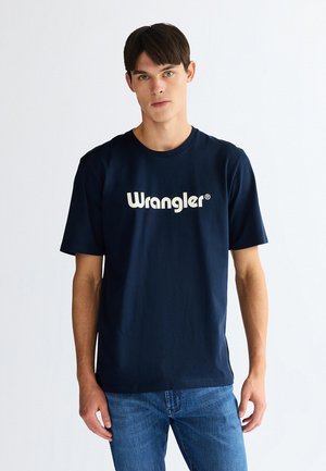 Junger Mann trägt ein dunkelblaues Wrangler T-Shirt und blaue Jeans, steht vor einem schlichten hellen Hintergrund und blickt nach vorne.