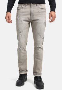 Jeans desgastados de color gris claro con rotos visibles y dobladillos deshilachados, con un diseño clásico de cinco bolsillos, combinados con zapatillas negras.