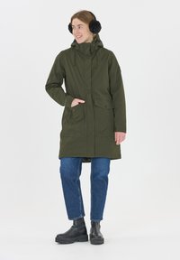 Groene waterdichte parka met een capuchon, zijzakken en verstelbare trekkoorden. Gerd gedragen over blauwe spijkerbroeken en zwarte stevige laarzen, tegen een witte achtergrond.