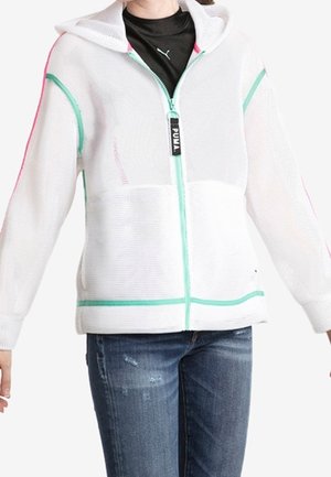 Persoon die een wit mesh hoodie met rits draagt, met turquoise en roze accenten en een blauwe spijkerbroek, staand met licht gespreide armen.