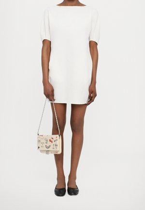 Vrouw in een wit mini-jurkje met korte mouwen, zwarte flats, en een beige handtas met zwarte en rode grafische prints op een kettingriem.