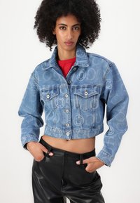 Veste en denim bleu raccourcie avec des motifs de smileys, fermeture à boutons, deux poches poitrine et un délavage. Associée à un pantalon en cuir noir.