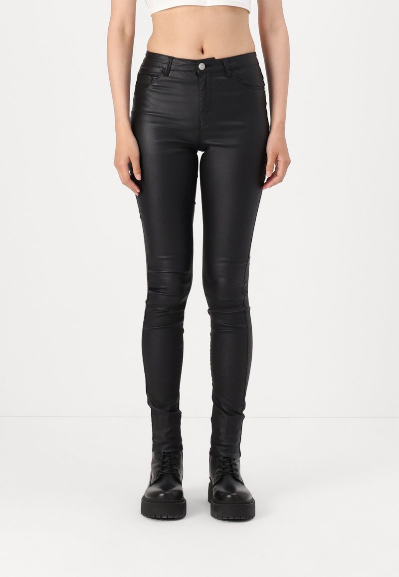 Pantalon en cuir noir coupe slim porté avec des bottes noires à lacets et un haut blanc court, visible de la taille aux pieds sur un fond neutre.