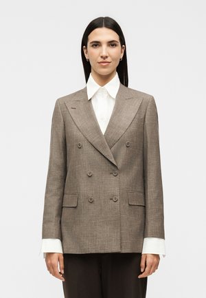 Sirgete tumedate juustega naine kannab valge kraega särgi peal pruuni kahekordse nööbitusega bleiserit ühevärvilise tausta ees.