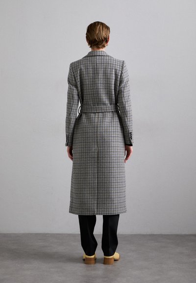 Long plaid coat in grey and black with a belt at the waist, long sleeves, and a tailored fit, paired with black pants and yellow shoes.