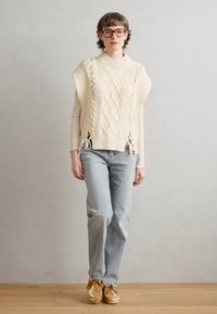 Maglione di crema chiara in maglia con motivi a treccia, vestibilità ampia e spacchi laterali, abbinato a jeans blu chiaro e sneakers beige.