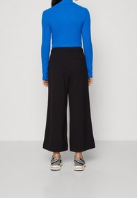 Blå långärmad topp, svarta vida byxor och grå sneakers. Byxorna har en slät textur med en bakfickadetalj.