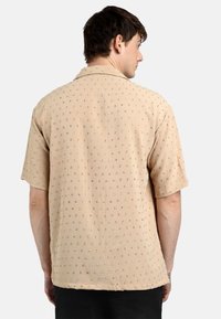 Camisa de manga corta beige con tejido texturizado que presenta patrones bordados coloridos, diseño de cuello y un ajuste relajado.