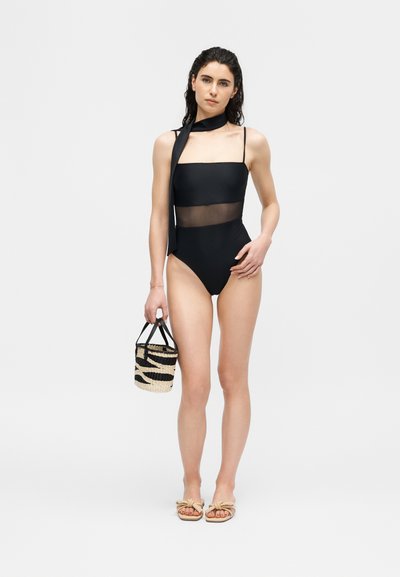 Femme portant un maillot de bain une pièce noir avec une partie transparente au milieu, tenant un petit sac en paille, et portant des sandales beige avec des nœuds.