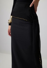 Gonna nera con vita strutturata, caratterizzata da borchie dorate lungo la parte superiore e un dettaglio drappeggiato con fiocco. Tessuto liscio e design lungo fino al pavimento.