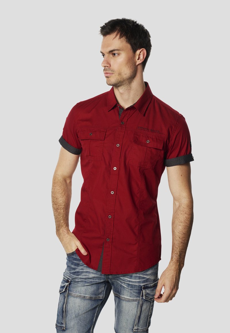 Camisa roja de manga corta con tapeta de botones y dos bolsillos en el pecho. Presenta detalles en denim oscuro en las mangas y el cuello. Se lleva con pantalones cargo vaqueros azules.